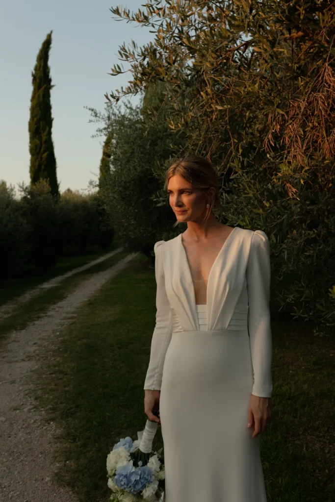 Home 41 A woman in a long-sleeved white wedding dress stands on a path beside green trees, holding a bouquet of flowers at her side, bathed in warm evening sunlight.