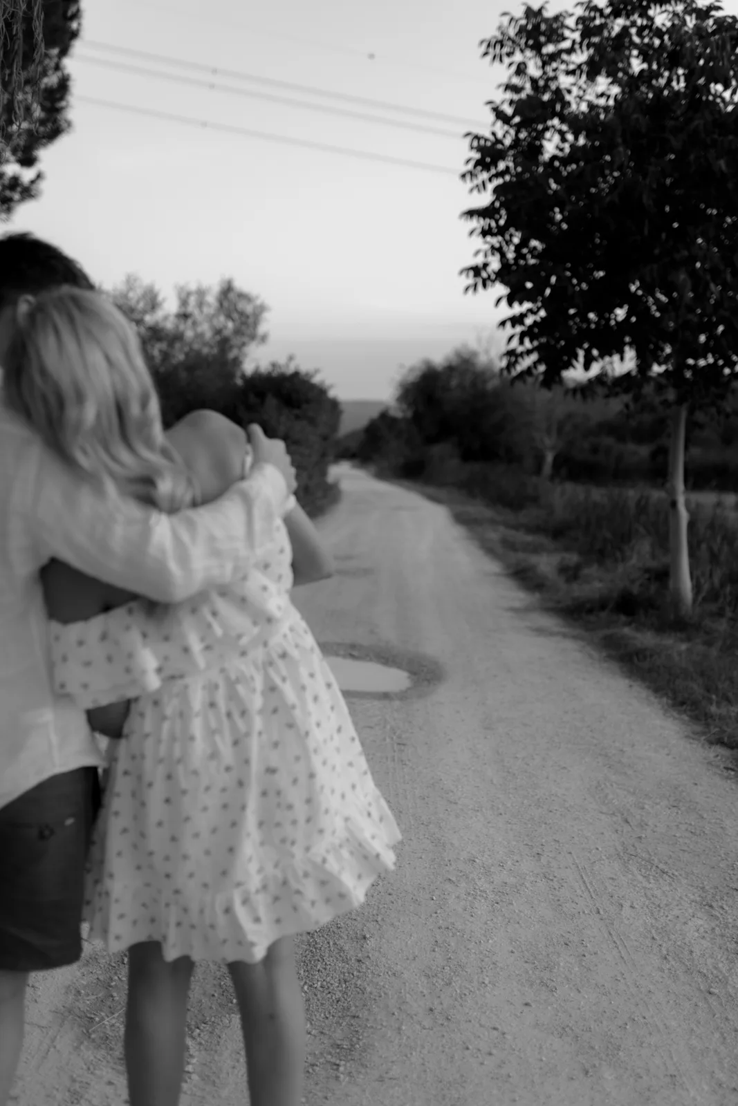Home 35 A black-and-white photo of two people embracing on a dirt road lined with trees, with one person wearing a light dress and the other in a shirt and shorts; the road stretches into the distance.