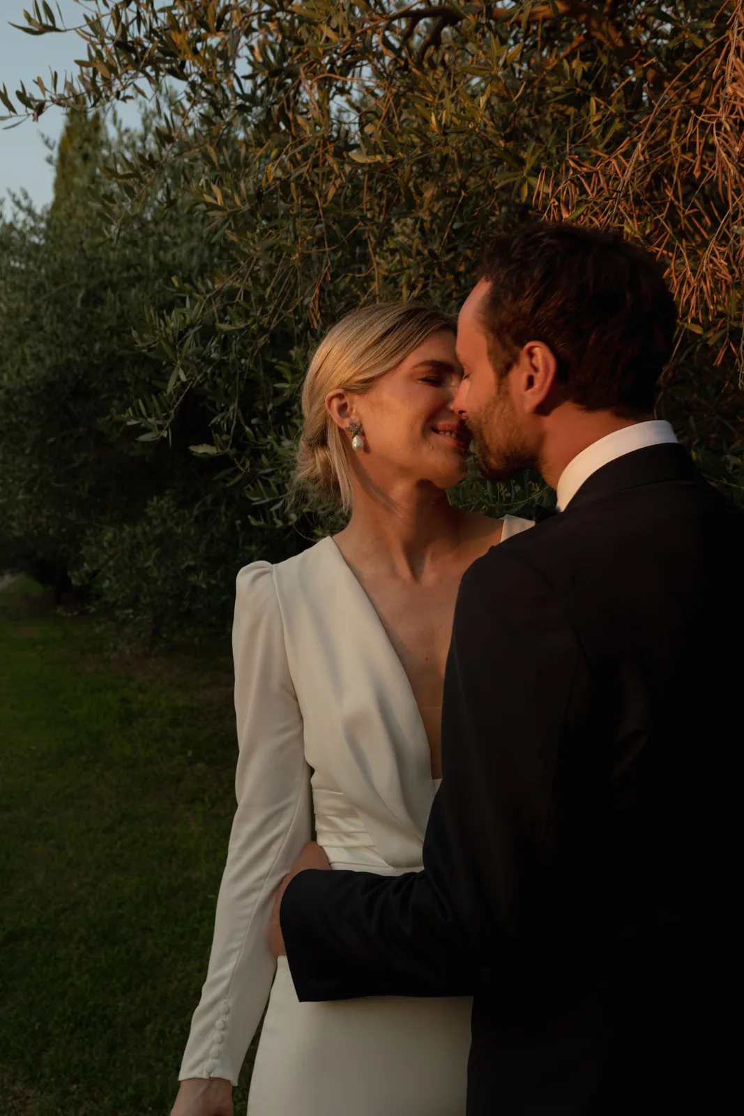 Home 24 A bride in a white dress and a groom in a black suit share a close, loving moment outdoors, surrounded by greenery and softly lit by warm sunlight.