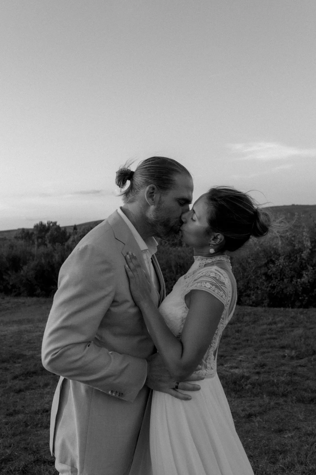 Kind Words 20 A bride and groom share a kiss outdoors at sunset. The bride wears a lace dress and the groom wears a light-colored suit. The background features trees and open sky, creating a romantic, peaceful scene.