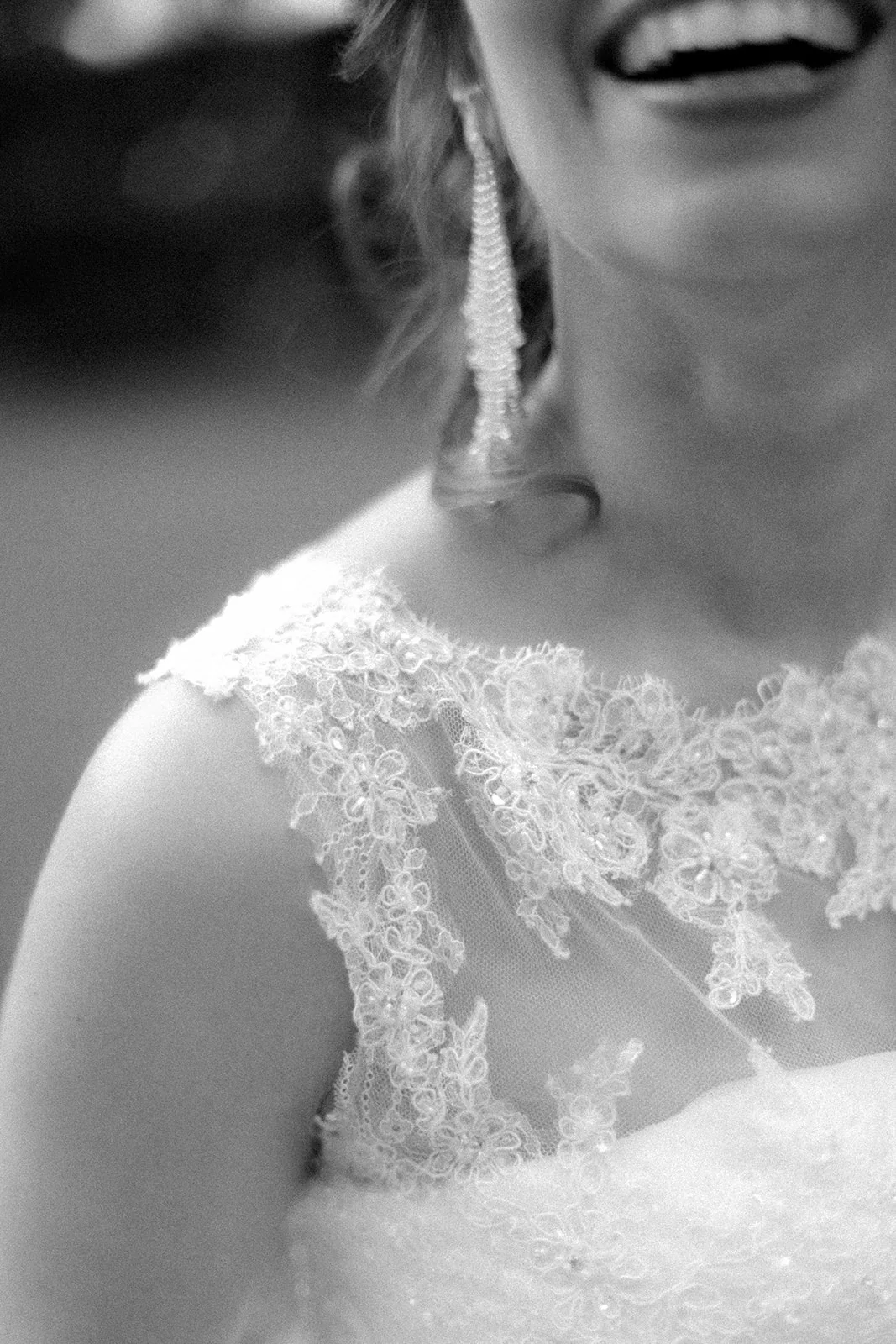 Home 30 Black and white close-up of a smiling bride wearing a lace wedding dress and long, sparkling earrings; only the lower half of her face and upper torso are visible.