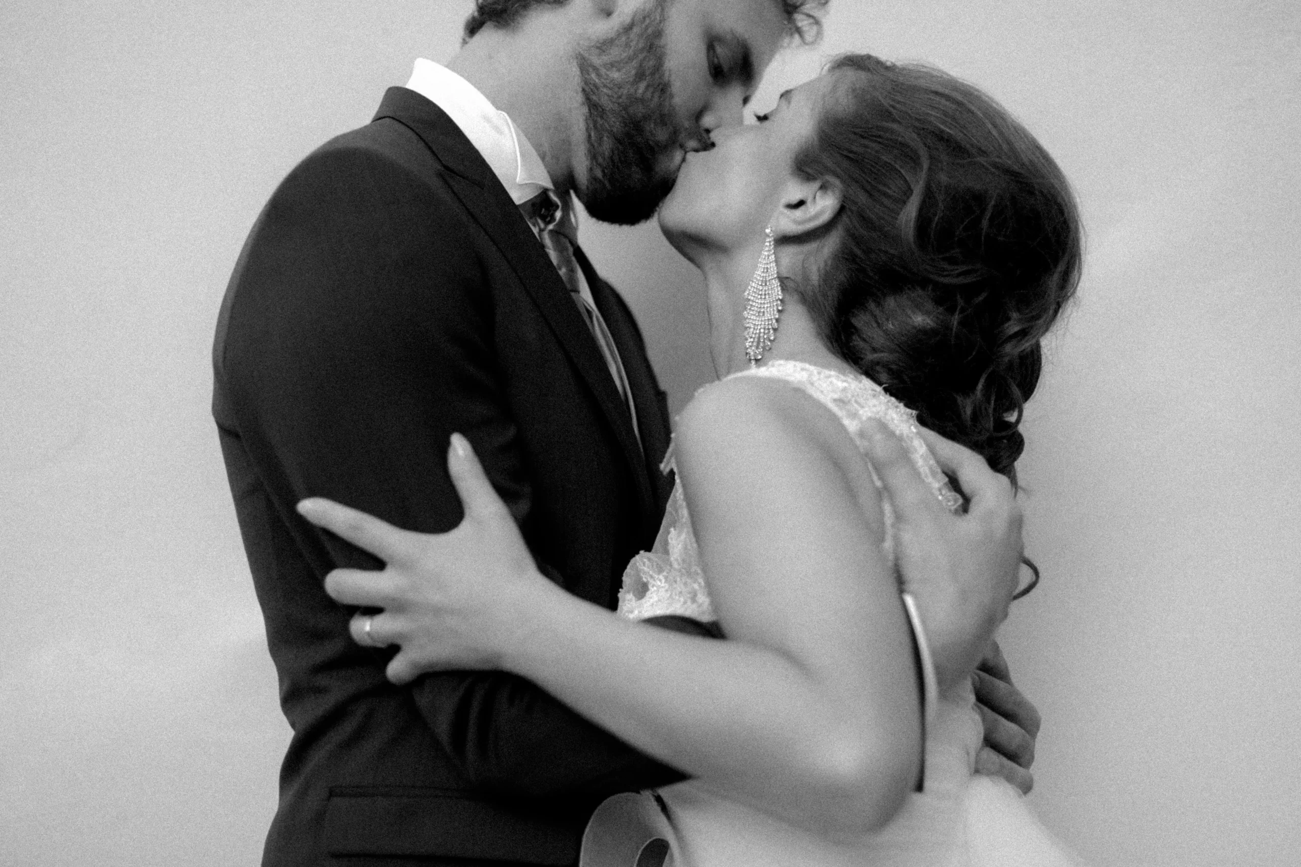 Kind Words 16 A bride and groom share a passionate kiss, dressed in wedding attire. Captured by an Italian wedding photographer, the black and white photo highlights the bride’s elegant gown and statement earrings as romance fills this intimate moment.