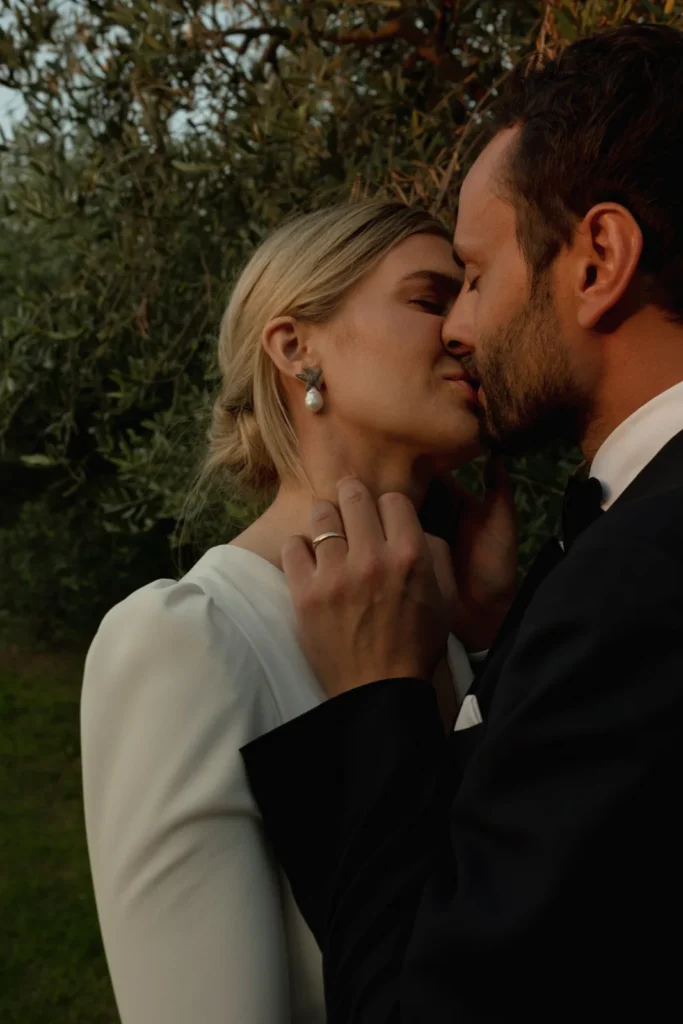 Home 12 A couple dressed in formal wedding attire shares an intimate moment, about to kiss, outdoors near green foliage. The woman wears a white dress and pearl earrings; the man wears a black suit and wedding ring.