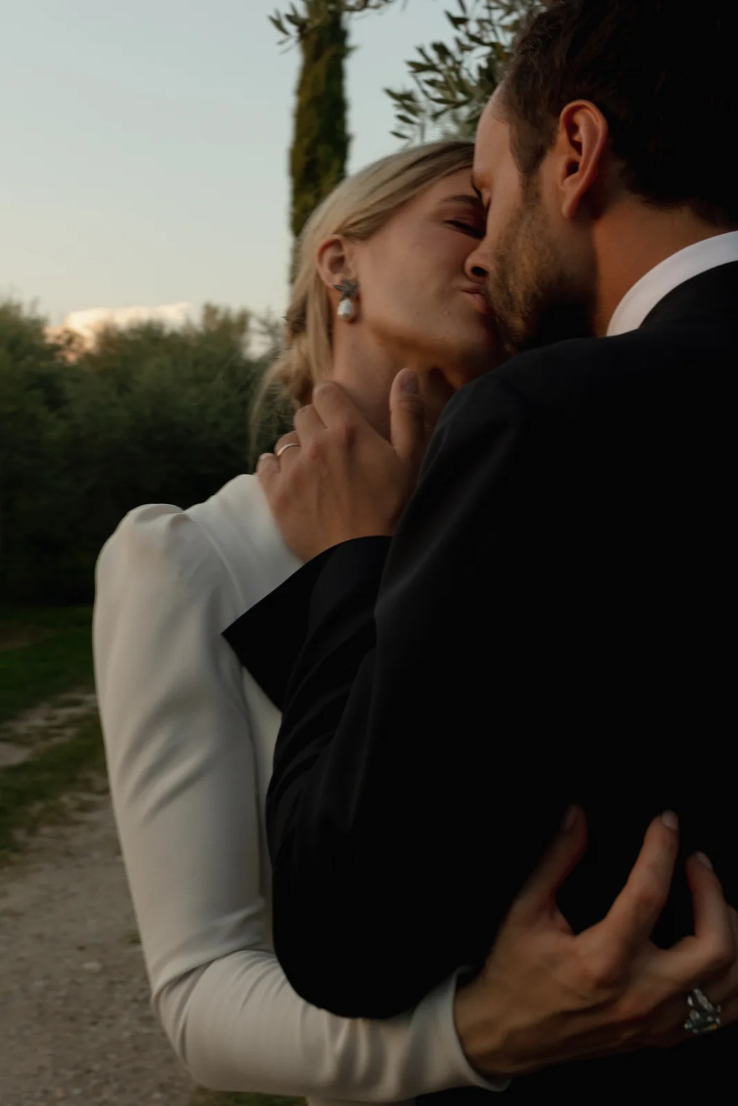 Home 32 A couple dressed formally, with the woman in white and the man in black, shares an intimate kiss outdoors on a path lined with greenery and trees at sunset.