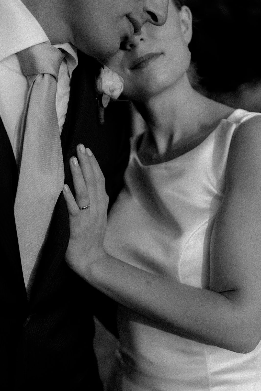 Home 17 A black and white photo of a bride in a satin dress resting her head on a groom’s chest. The groom wears a suit and tie, and the bride’s hand with a wedding ring rests on his shoulder.