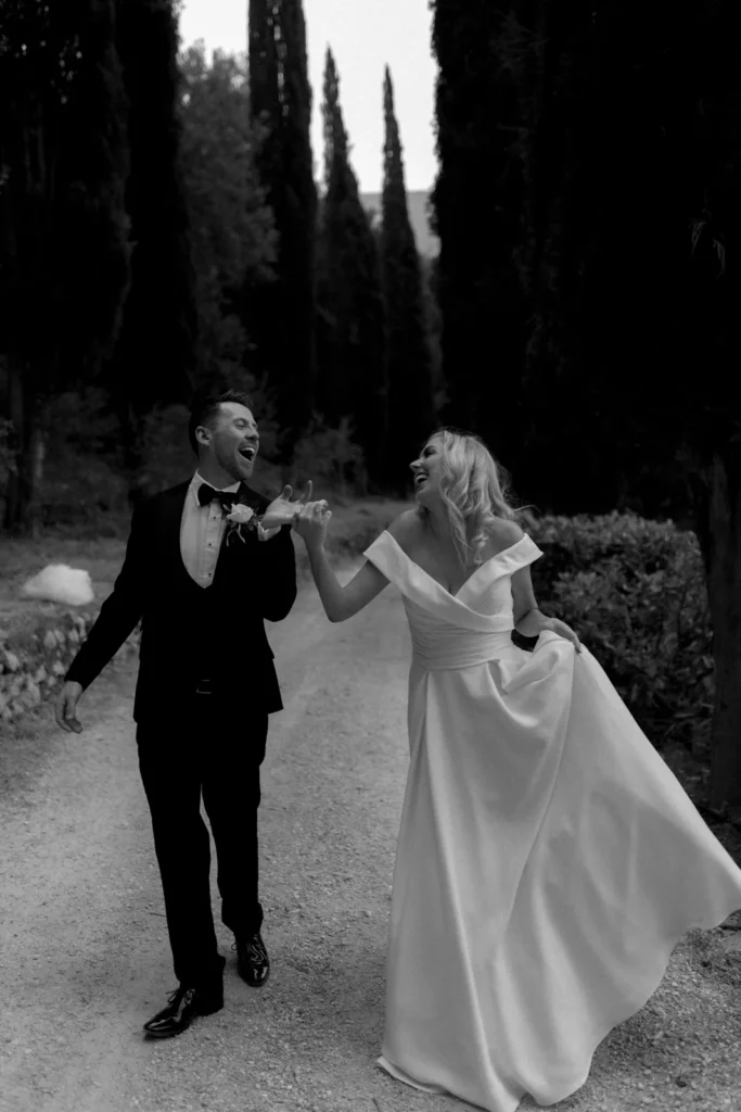 Home 11 A joyful bride and groom walk hand in hand down a tree-lined path, smiling and laughing together. The bride wears an off-shoulder gown and the groom is in a tuxedo. The scene is in black and white.