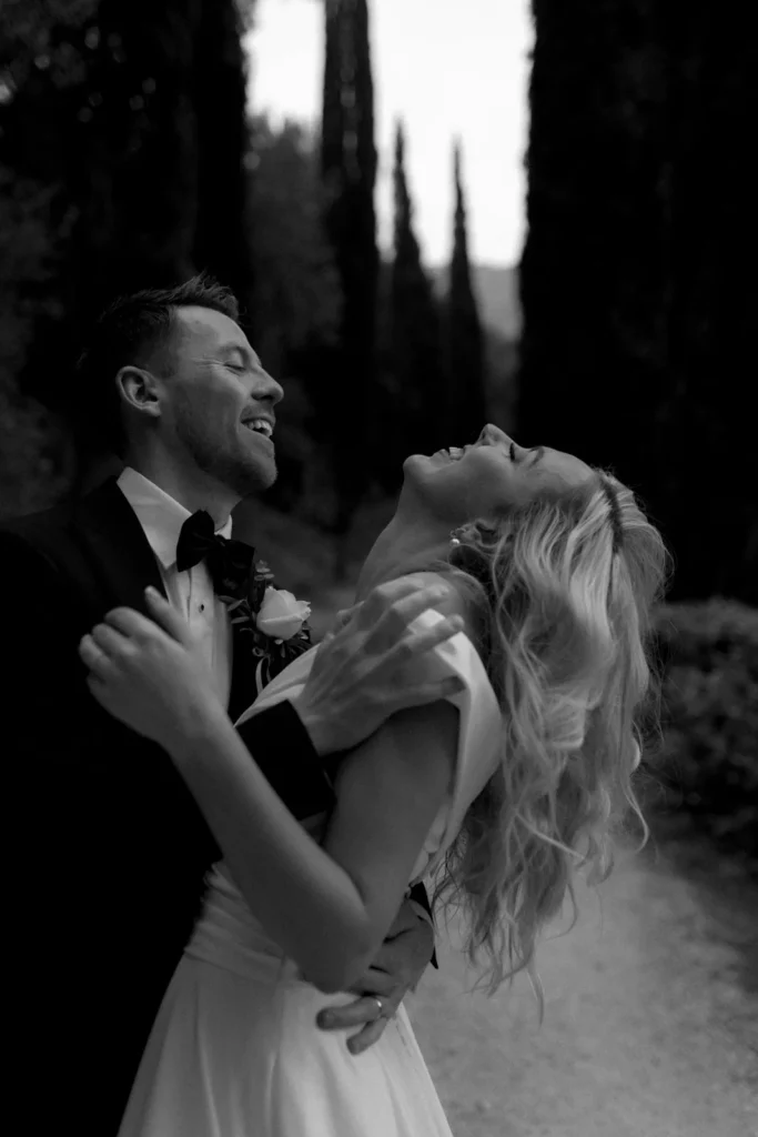 A groom in a tuxedo embraces a bride in a white dress, both laughing joyfully outdoors.