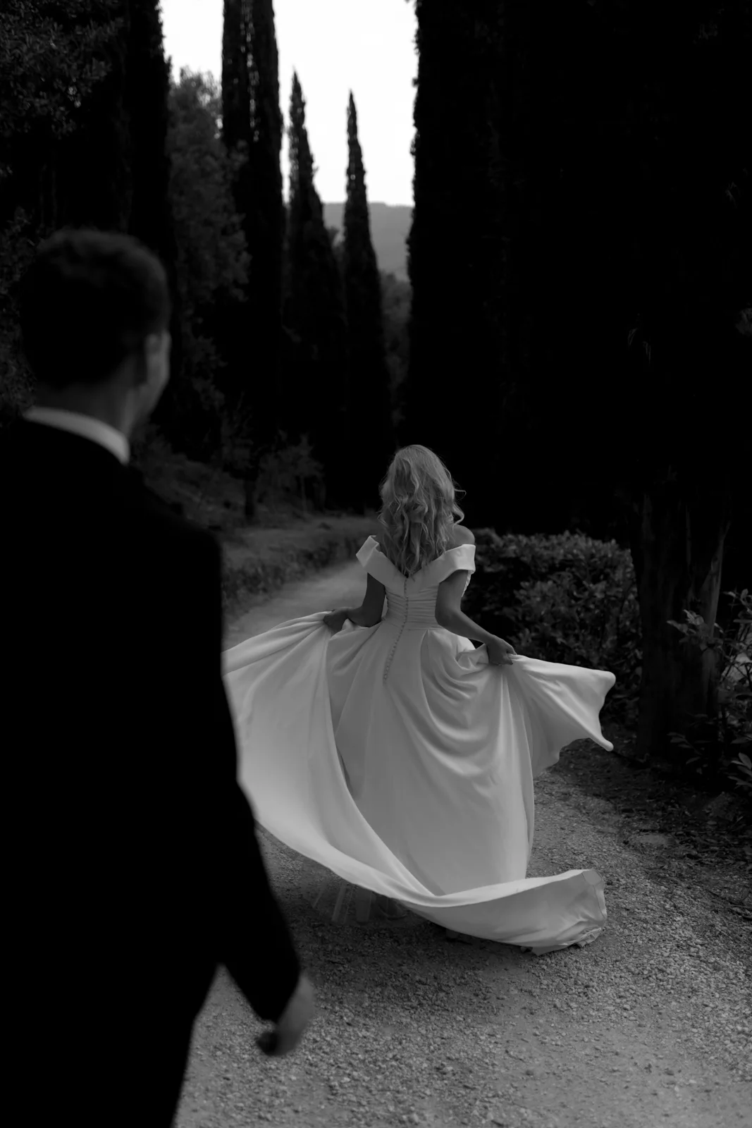 Home 29 A black-and-white photo of a woman in a flowing white dress walking away on a path lined with tall trees, while a man in a dark suit watches her from behind.