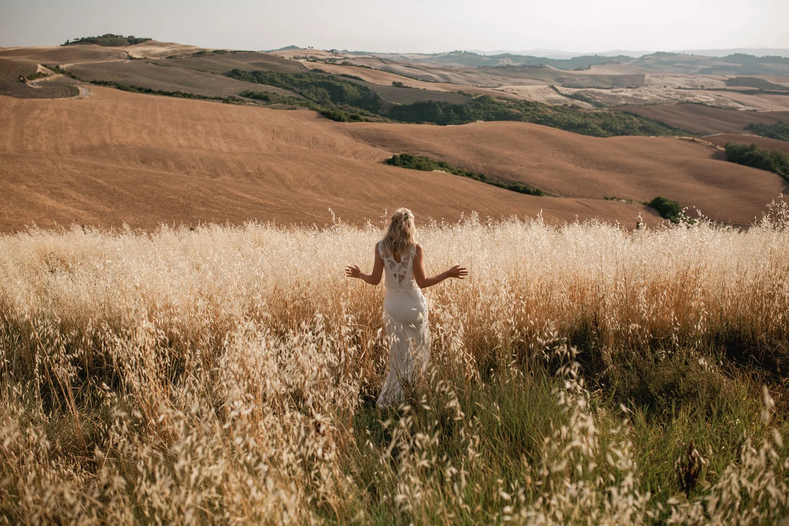 A woman in a white dress stands in tall, golden grass, facing away from the camera, as a Tuscan wedding photographer captures her overlooking rolling brown hills and fields beneath a bright sky.