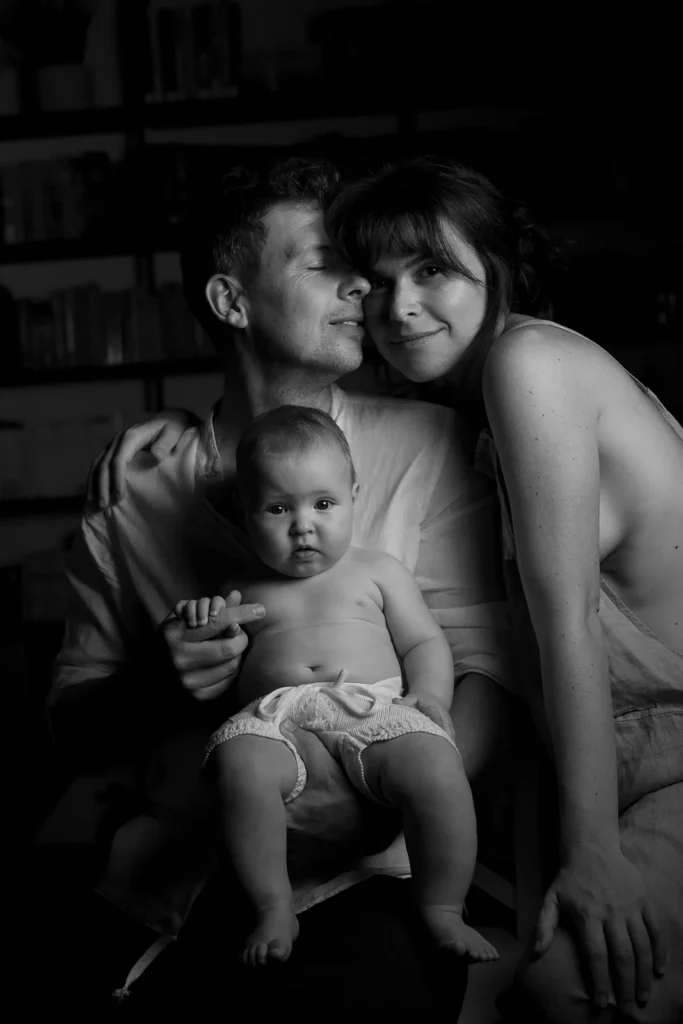 A black-and-white photo by a wedding photographer in Italy shows a couple sitting closely, smiling, with a baby in the man's lap. The baby looks at the camera while the parents share gentle, affectionate expressions.