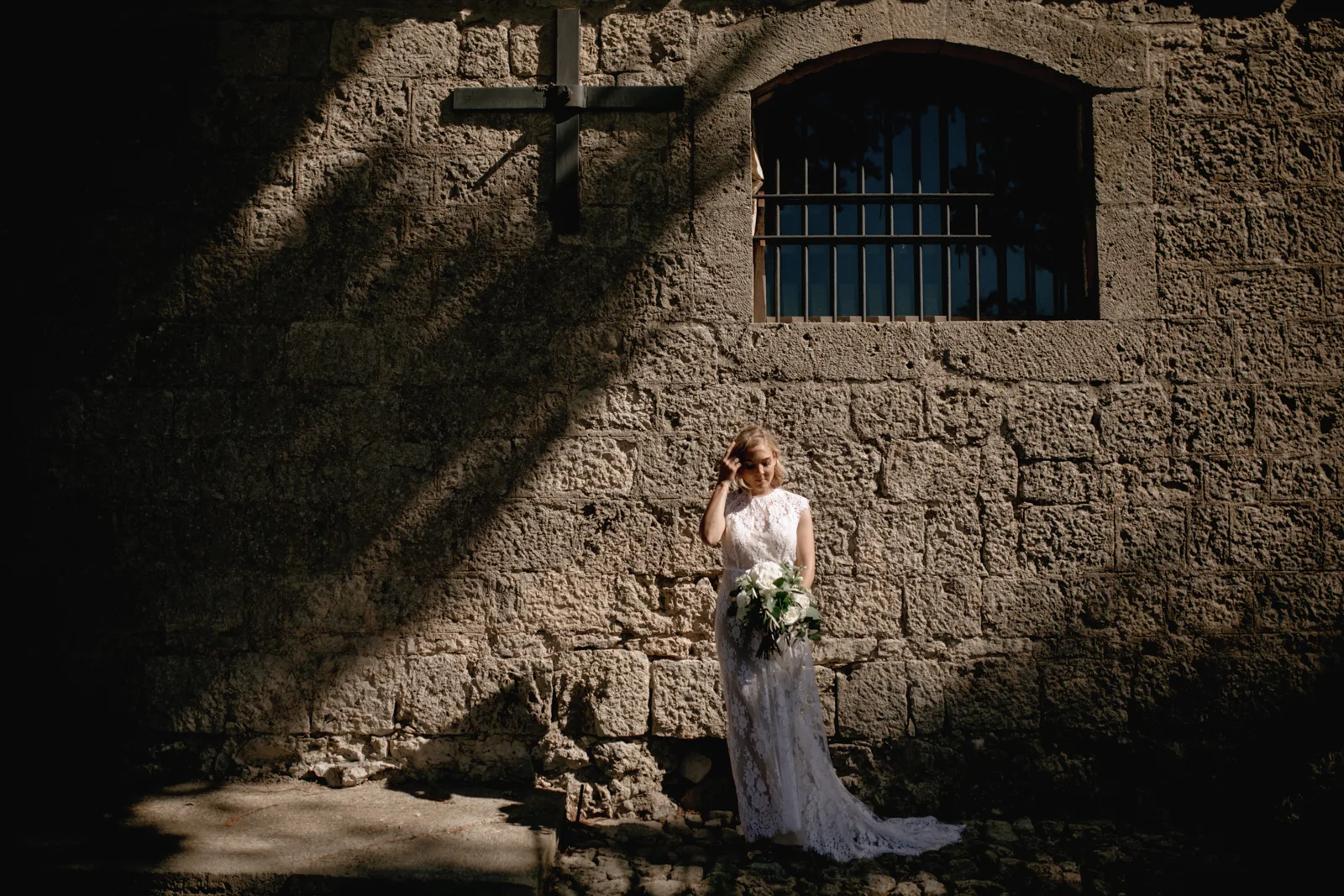 Home 34 A bride in a white lace gown stands alone against a textured stone wall in sunlight and shadow, holding a bouquet of flowers. A barred window and a cross-shaped shadow appear above her on the wall.