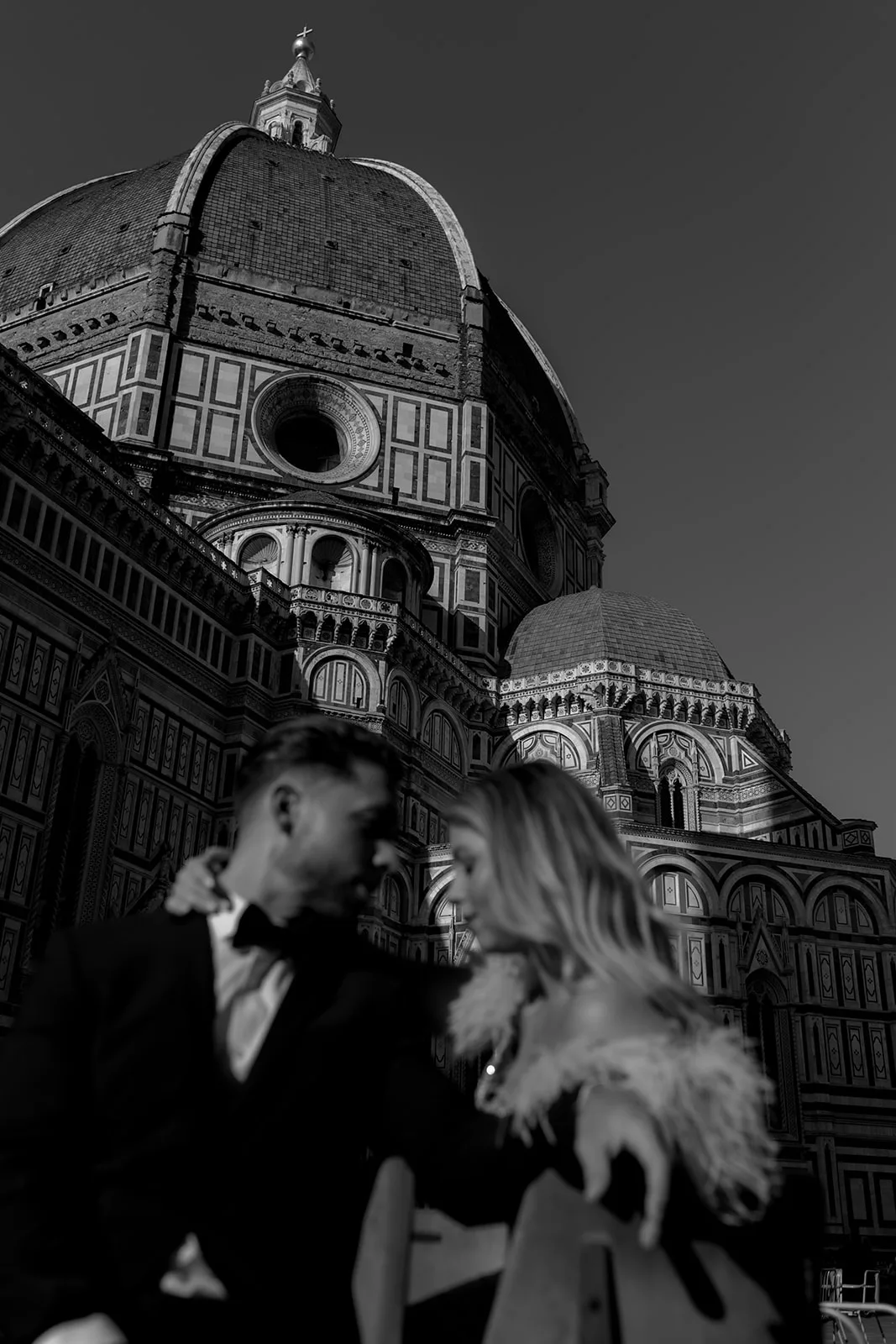 Home 28 A couple dressed elegantly sits closely together in front of the Florence Cathedral dome, with the historic architecture rising dramatically behind them in black and white—perfectly captured by a Tuscan wedding photographer.