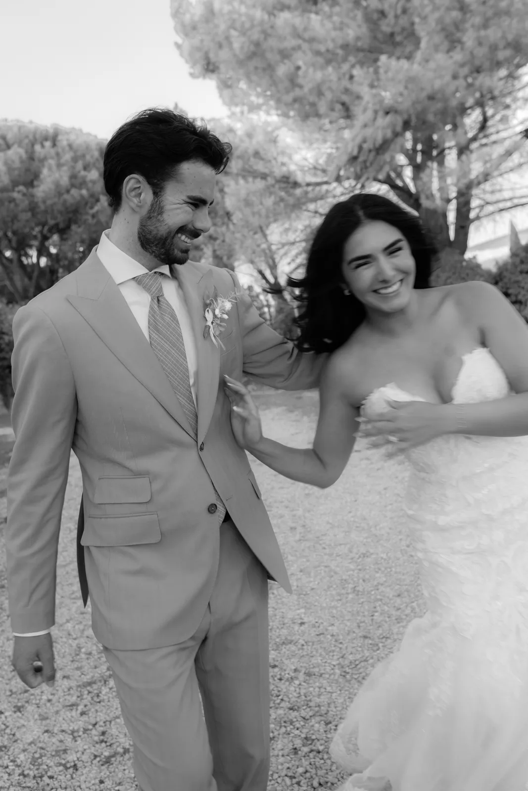Home 45 A joyful bride and groom, captured by an Italian wedding photographer, walk hand in hand outdoors, smiling and laughing together. The black and white photo features the happy couple with trees beautifully framing the moment in the background.