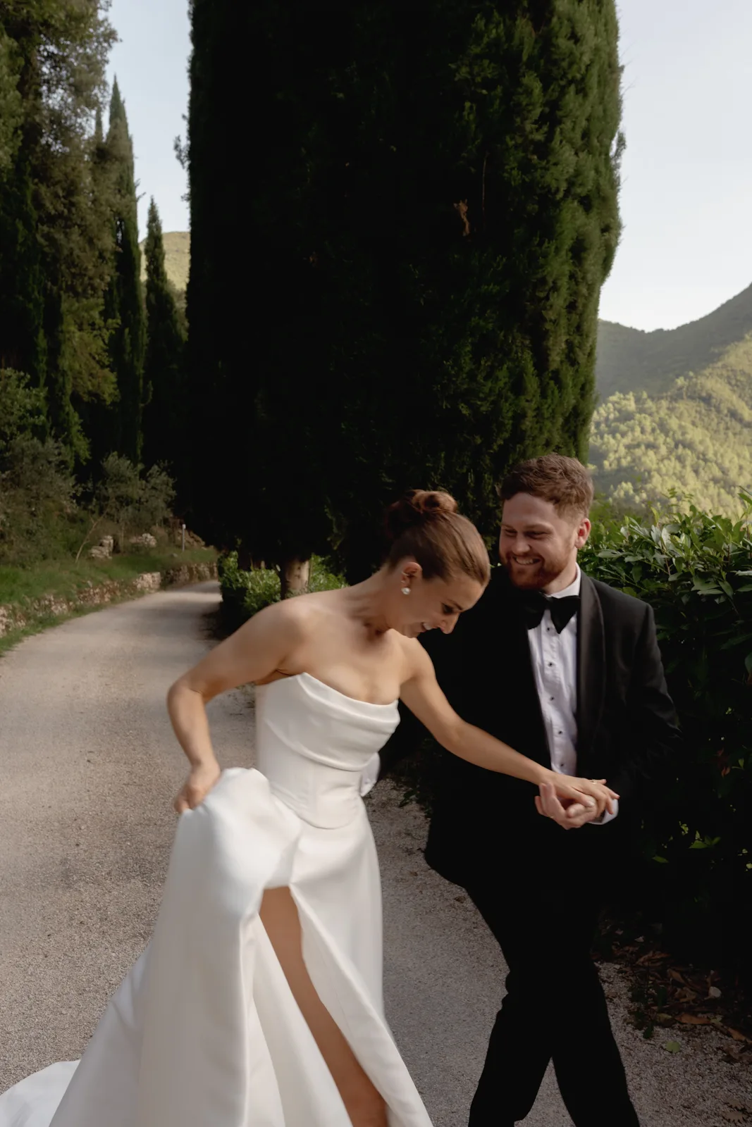 Home 46 A bride in a white strapless gown holds up her dress while playfully walking with a groom in a black tuxedo, beautifully captured by an Italian wedding photographer on a scenic path lined with greenery and mountains in the background.