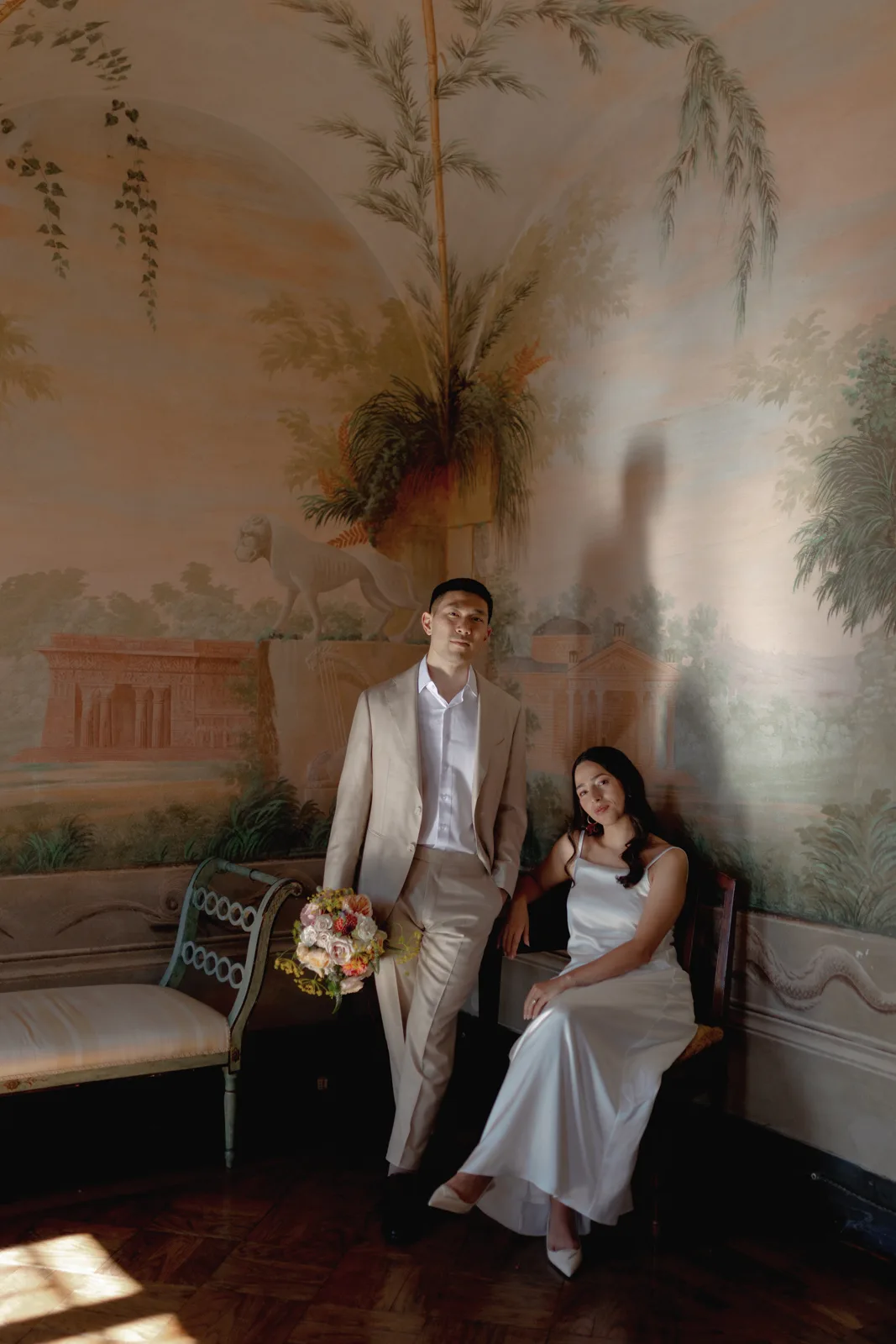 Home 44 A couple in formal attire poses in a softly lit, ornately painted room. Captured by an Italian wedding photographer, the man in a beige suit stands beside a seated woman in a white dress holding a bouquet, their shadows on the mural-covered wall behind them.