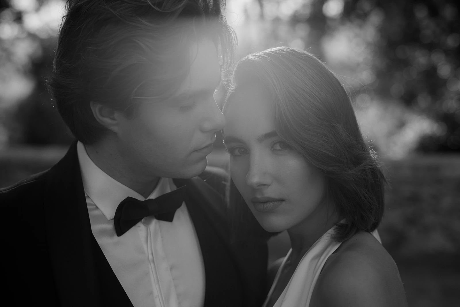 A black-and-white photo of a man in a tuxedo and a woman in a sleeveless dress, standing close together outdoors. The man looks at the woman while she gazes directly at the camera, sunlight softly illuminating them.