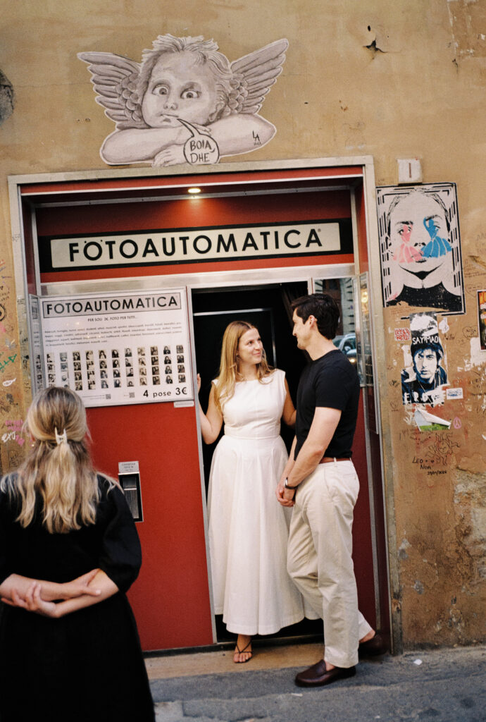 A smiling couple stands in the doorway of a vintage red photo booth labeled Fotoautomatica on a graffiti-covered street, while a woman with blonde hair stands facing them, back to the camera.