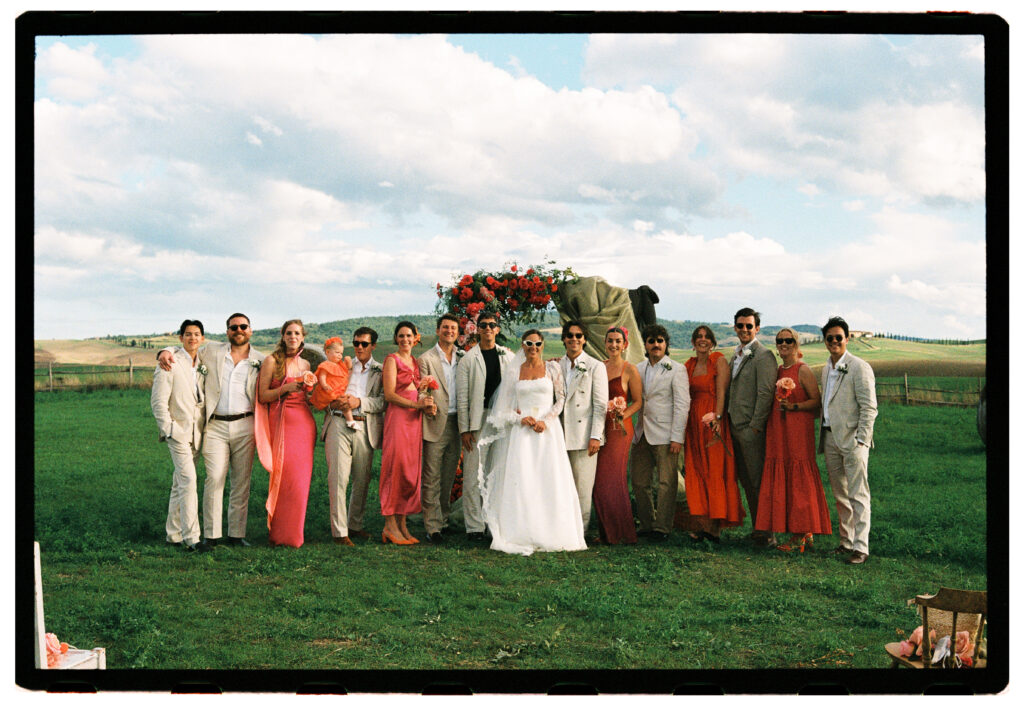 A wedding party of 16 people stands in a row on green grass, with the bride and groom at the center. Bridesmaids wear pink and coral dresses; groomsmen wear beige suits. A floral arrangement is visible behind them, with a blue sky overhead.