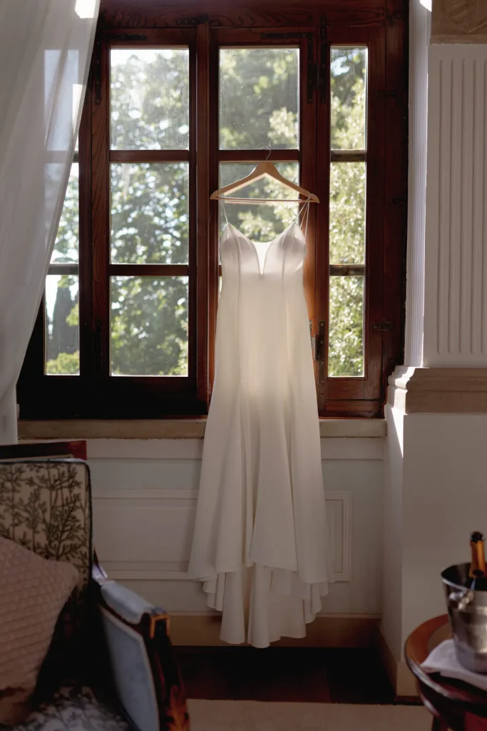 A white sleeveless wedding dress on a hanger is displayed in front of a sunlit wooden window, with greenery visible outside.