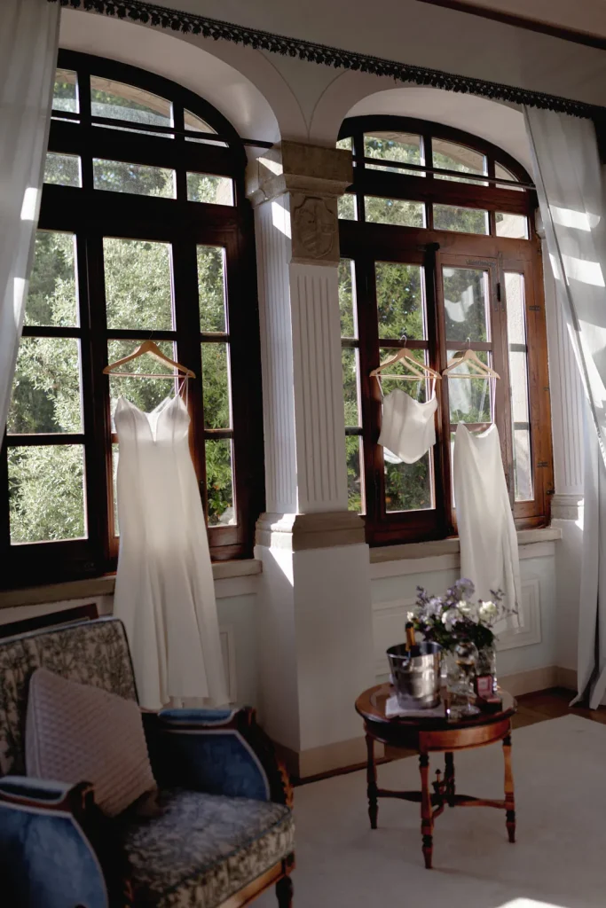 Two white dresses and a camisole on hangers are displayed in front of tall windows, with sunlight streaming in. A blue armchair and a round table with flowers and drinks are in the foreground.