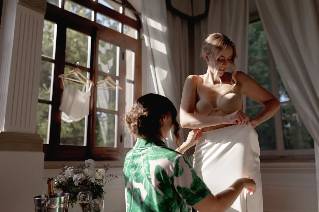 A woman helps another woman adjust her white skirt in a sunlit room with large windows, flowers, and a hanging white top in the background.