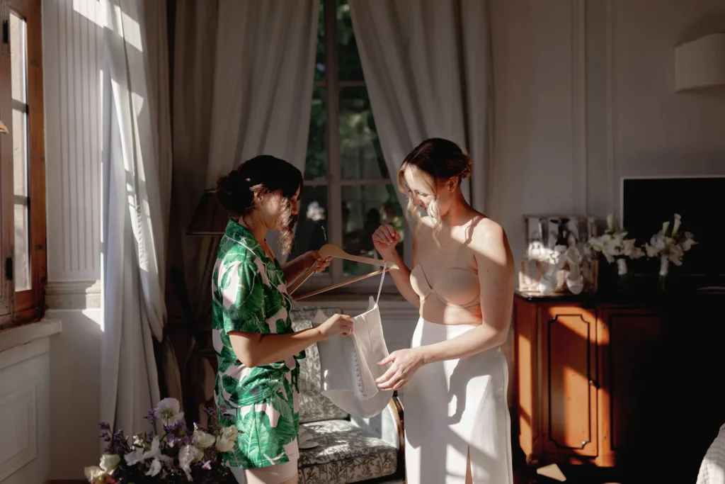 Two women stand by a sunlit window in a room with flowers and curtains. One holds a hanger with white lingerie; the other, in a bra and skirt, looks at it, both smiling in a relaxed, intimate atmosphere.