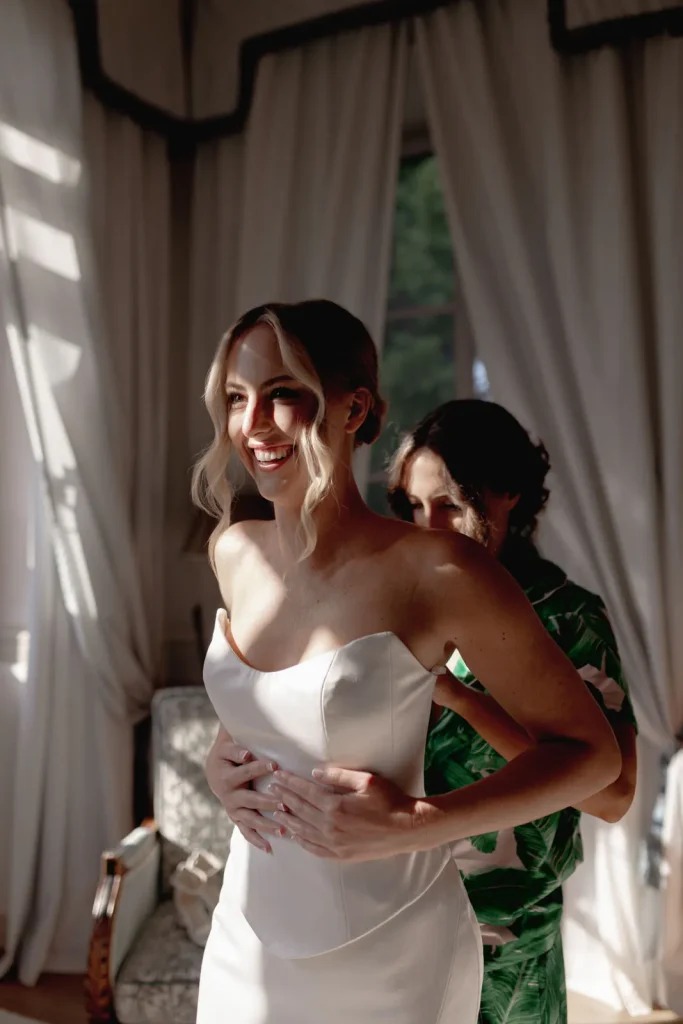 A smiling bride in a white dress stands as another woman helps her zip up the gown in a sunlit room with white curtains.