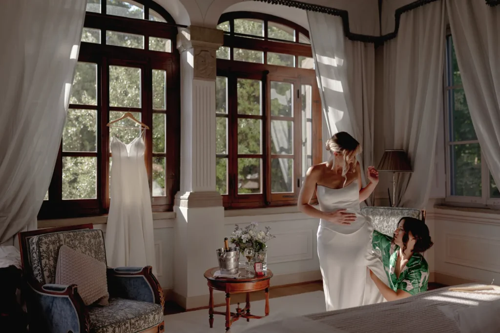 A woman in a wedding dress stands near a window while another woman in green helps adjust the dress. Sunlight streams in, and a second white dress hangs nearby in a bright, elegant room.