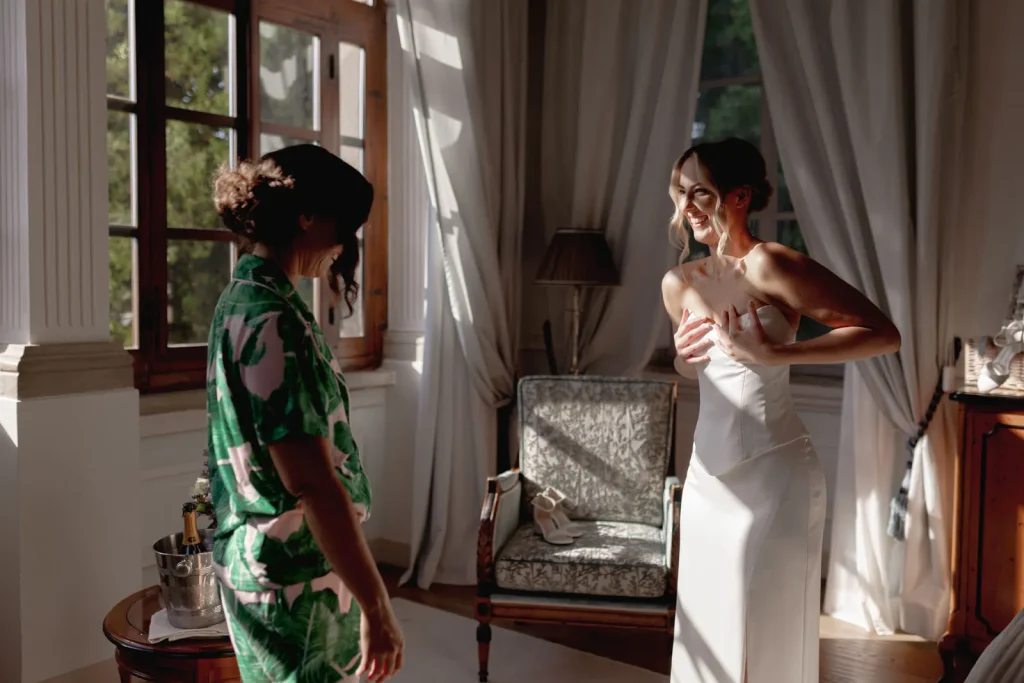 Two women in a sunlit room; one wears a green outfit, the other adjusts her strapless white dress while smiling. An armchair, curtains, and large window appear in the background.