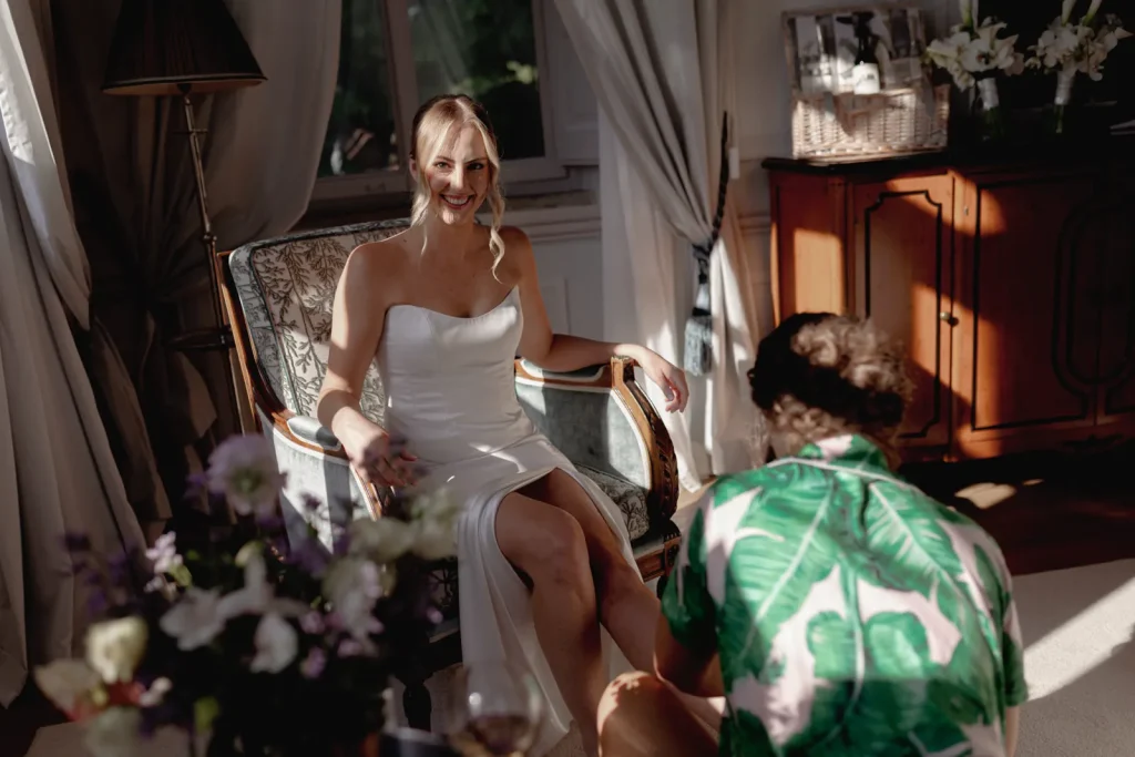 A woman in a white strapless dress sits smiling on a chair, while a person in a green leaf-patterned shirt kneels in front of her. Sunlight streams into the elegant room decorated with flowers.