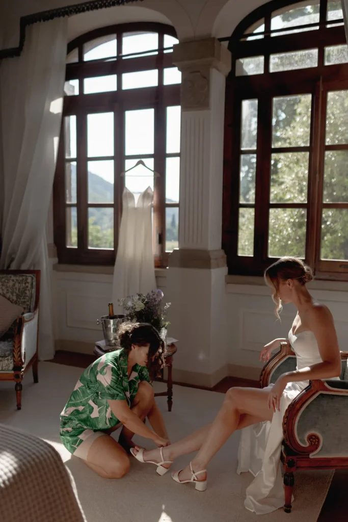 A woman in a white dress sits as another woman in green helps her put on shoes in a sunlit room with large windows. A white gown hangs in the background.