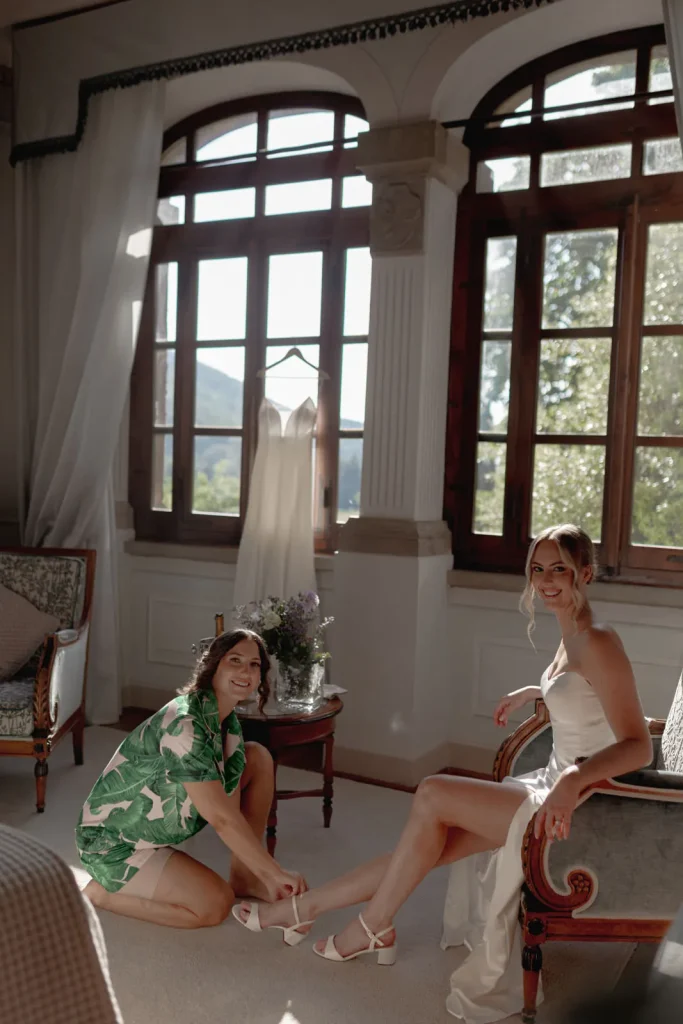 Two women are in a sunlit room. One, in a white dress, sits on a chair smiling, while the other kneels, adjusting her shoe. A wedding dress hangs in the background by large windows.