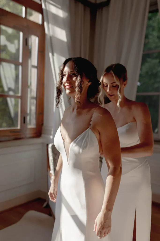 Two women in white dresses stand by sunlit windows; one smiles as the other fastens the back of her dress, suggesting a bridal preparation scene in a softly lit room with curtains and greenery outside.
