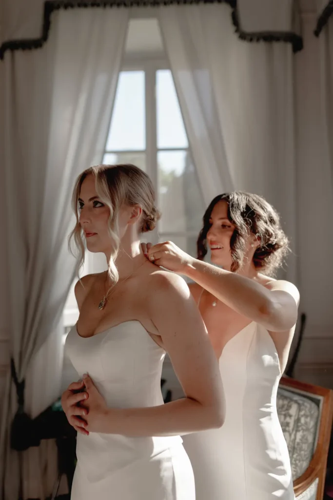 Two women in white dresses stand by a sunlit window. One woman helps fasten the other’s necklace. Both appear calm and focused, with soft light highlighting their features, suggesting a wedding or formal occasion.