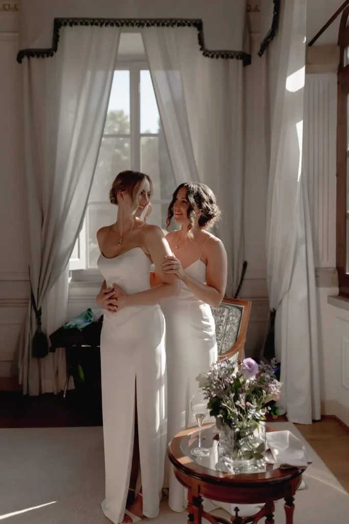 Two women in elegant white dresses embrace and smile warmly in a sunlit room with large windows and sheer curtains. A table with a floral arrangement is in the foreground.