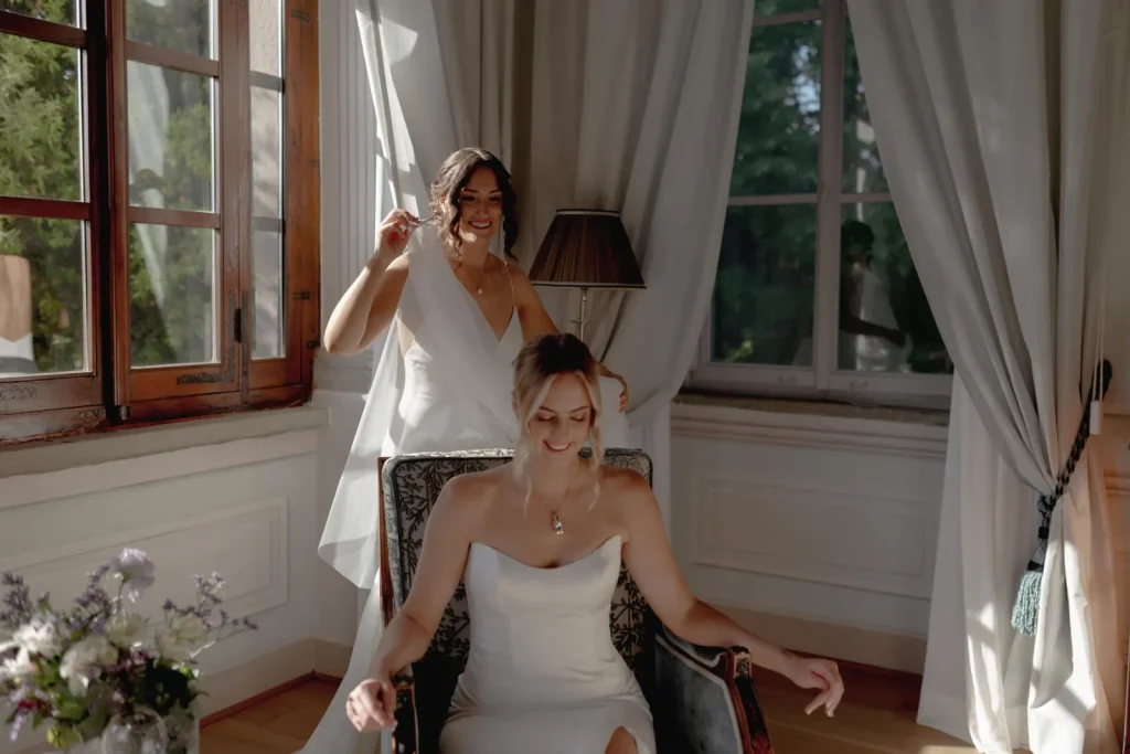 Two women in white dresses smile in a bright, elegant room. One sits on a chair, the other stands behind her, holding a necklace. Sunlight streams through large windows with white curtains. Flowers are visible on a table.