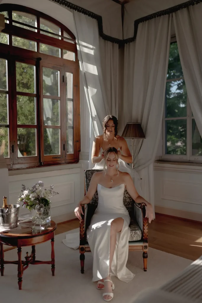 A bride in a white dress sits in an armchair, smiling, while another woman stands behind her fixing her hair. Sunlight streams through large windows, illuminating the elegant, softly decorated room.