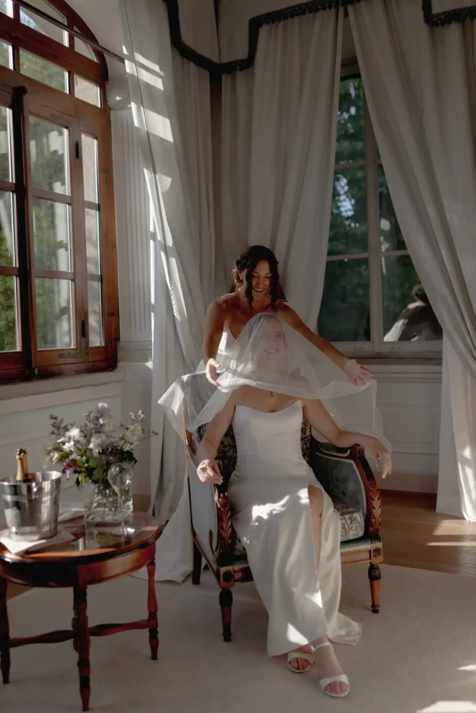 A bride sits on a chair in a sunlit room as another woman gently arranges her veil. A small table with flowers and a champagne bucket is nearby. Elegant curtains and large windows frame the scene.