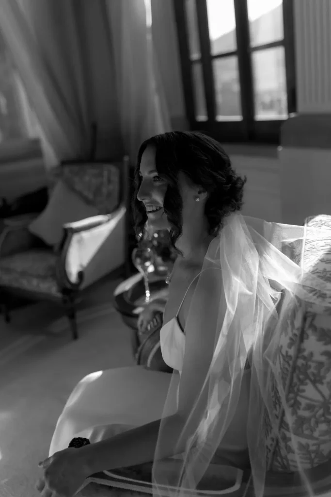 A bride in a white dress and veil sits on a chair in a sunlit room, smiling and looking to the side. The black and white photo captures elegant, vintage-style decor in the background.