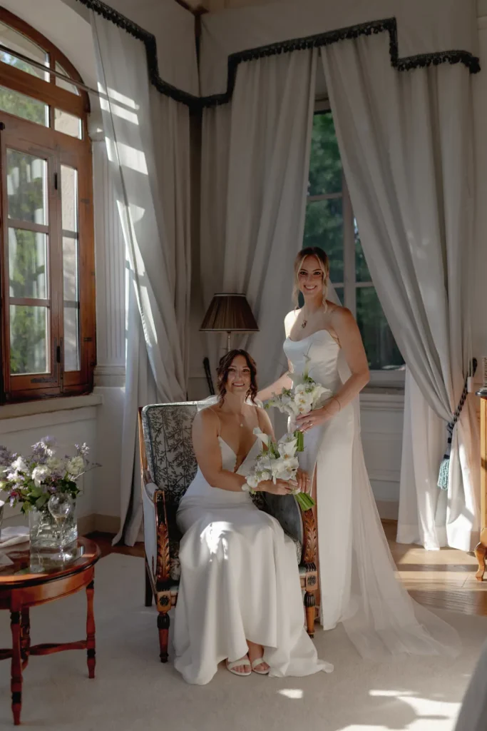 Two women in white dresses pose in a bright, elegant room with large windows and curtains. One woman sits holding a bouquet, while the other stands beside her, both smiling. A small table with flowers is nearby.