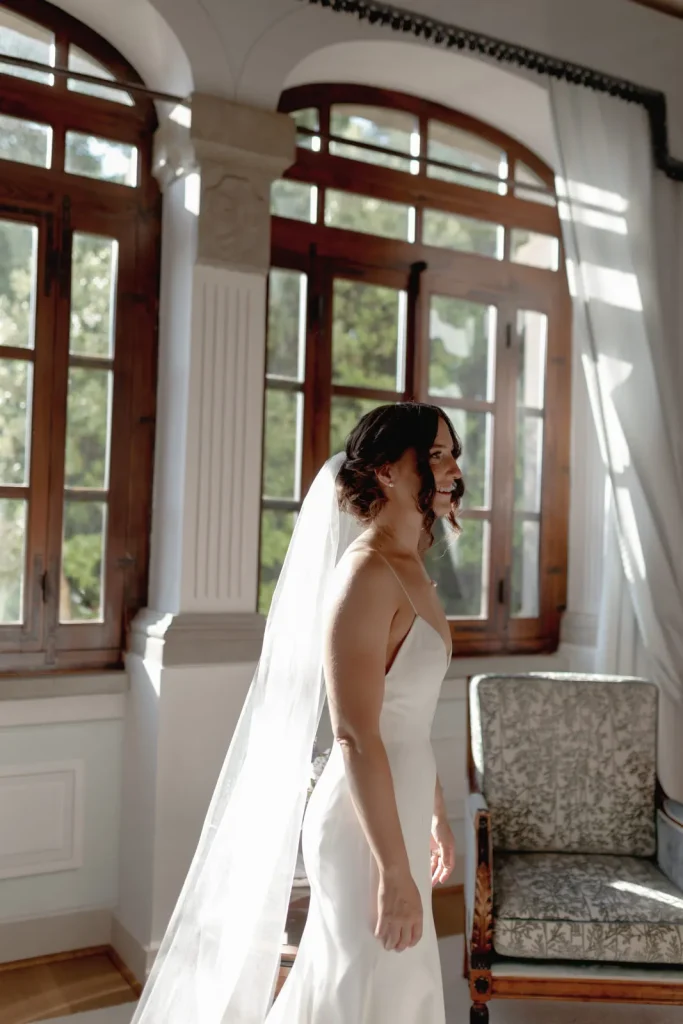 A bride in a white dress and veil stands by large, sunlit windows, smiling slightly. Ornate wooden frames and a patterned armchair are visible in the elegant room.