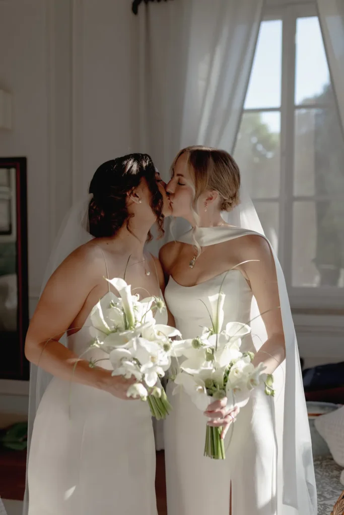 Two brides in white dresses and veils kiss while holding white flower bouquets in a sunlit room with a large window behind them.
