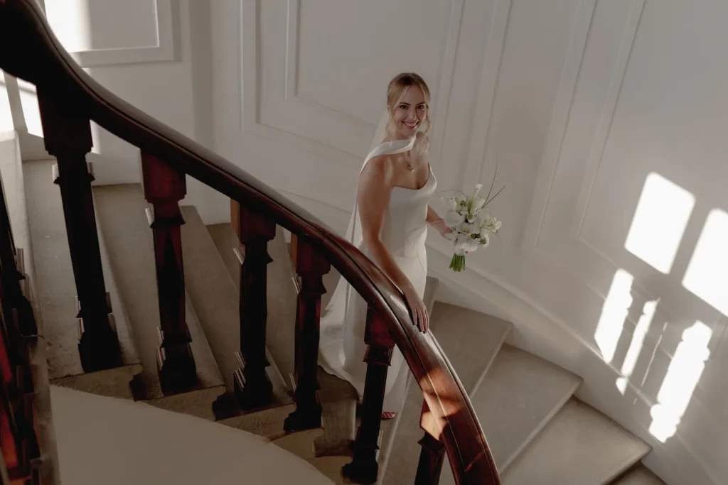 A bride in a white dress and veil holds a bouquet of flowers while standing on a curved staircase, smiling up toward the camera. Sunlight streams through windows, casting shadows on the wall.