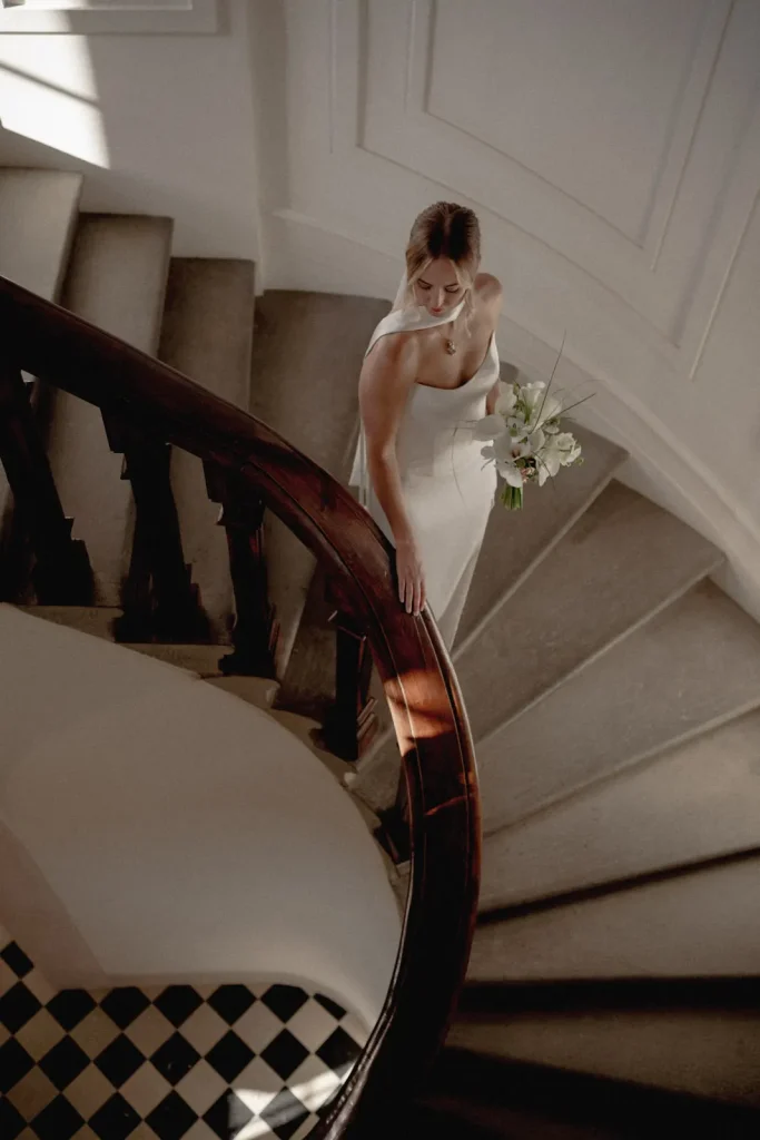 A bride in a white dress holds a bouquet while walking down a curved staircase with a dark wooden railing and checkered floor below.