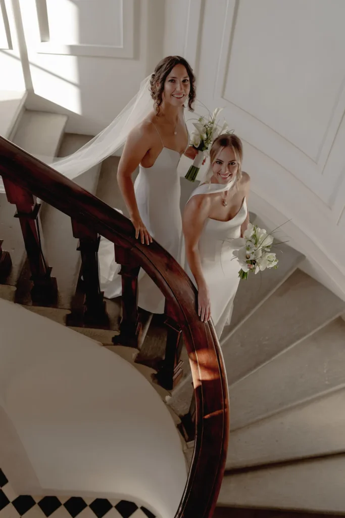 Two women in white dresses holding bouquets and smiling stand on a curved staircase with elegant white walls and soft sunlight coming through a window.