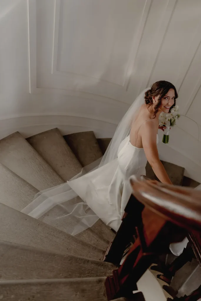 A bride in a white dress and veil holds a bouquet while walking up a spiral staircase, looking back over her shoulder and smiling.