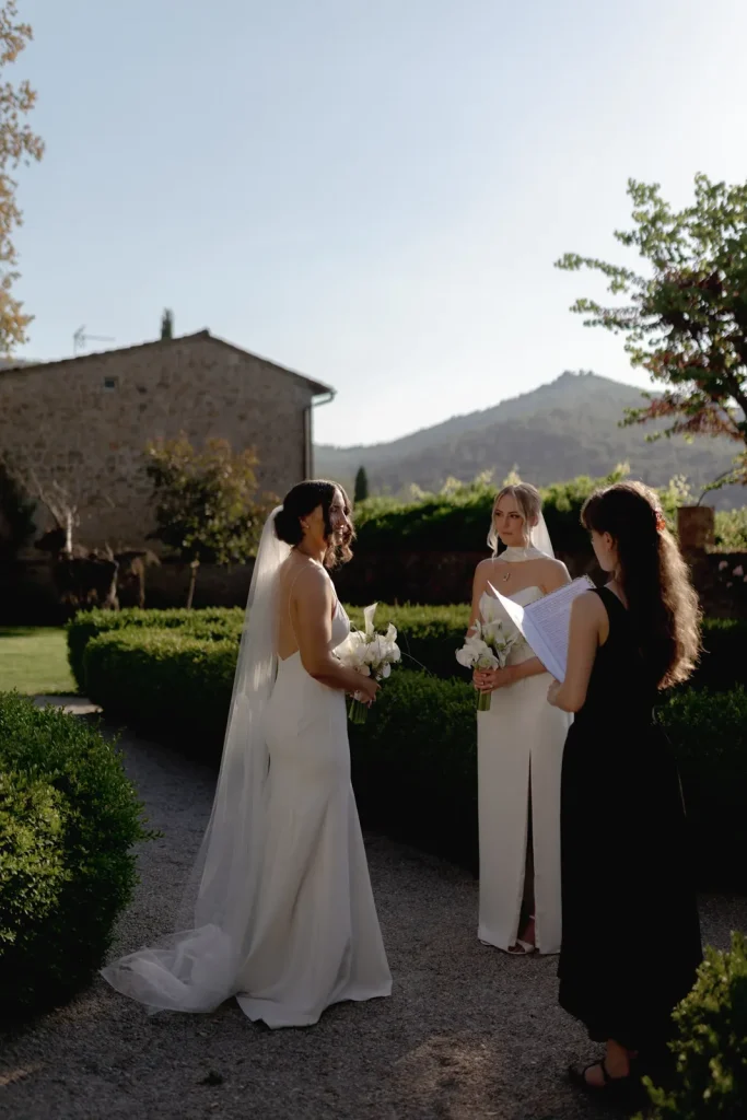 A bride in a white dress stands with her bridesmaid and an officiant outdoors, surrounded by greenery and stone buildings, during a wedding ceremony on a sunny day.