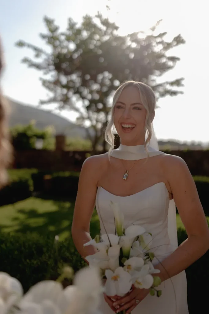 A bride in a strapless white dress holding a bouquet of white flowers smiles outdoors in sunlight, with greenery and a tree in the background.
