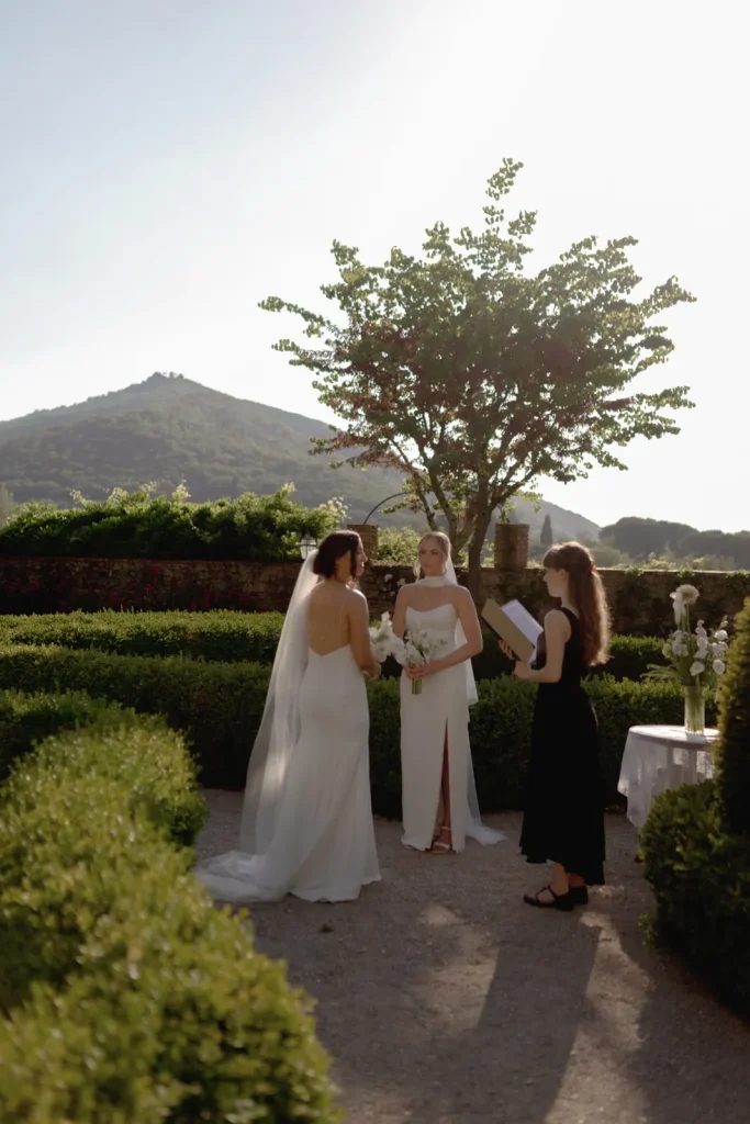 Two brides in white dresses stand together during an outdoor wedding ceremony, facing an officiant with a book. Green hedges and a tree surround them, with a sunlit mountain in the background.
