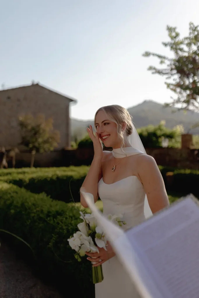 A bride in a white dress and veil smiles and wipes a tear from her face while holding a bouquet, standing outdoors in a garden with sunlight and hills in the background.